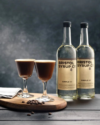 Two bottles of Bristol Syrup Co. on a wooden board with two glasses of coffee.