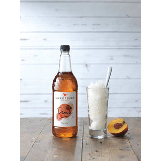 Bottle of Sweetbird Peach syrup with a glass of ice and a peach on a wooden surface.
