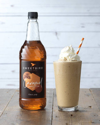 Bottle of Sweetbird Caramel syrup next to a glass of a caramel-themed drink with whipped cream and a straw on a wooden surface.