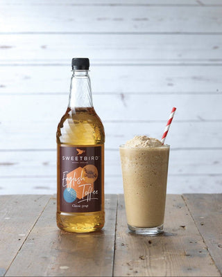 Bottle of Sweetbird English Toffee syrup next to a glass of iced frappe with a straw on a wooden surface.