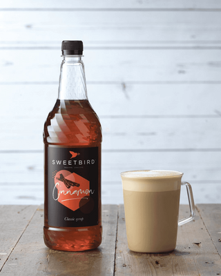 Bottle of Sweetbird cinnamon syrup next to a cup of cinnamon latte on a wooden surface.